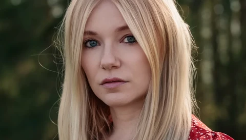 Portrait of a woman with platinum blonde hair and blue eyes against a blurred forest background.