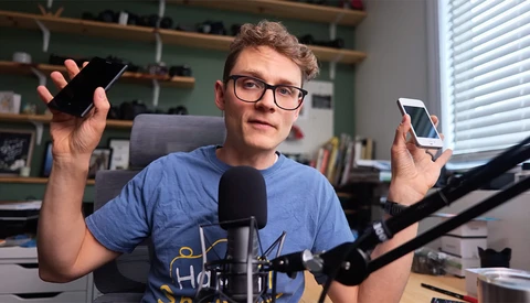 Young content creator holding multiple smartphones while speaking into a microphone at a desk setup.