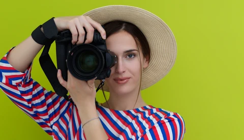 Woman in striped shirt and straw hat holding DSLR camera up to her face against yellow background.