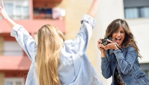 Young woman photographing another woman with arms raised in celebration outdoors.
