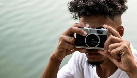 Photographer holding a rangefinder camera up to his face against a blurred water background.