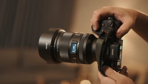 Hands holding a mirrorless camera with attached lens against a warm, blurred background.