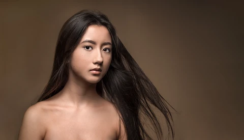 Woman with long black hair against neutral brown background, looking toward camera.