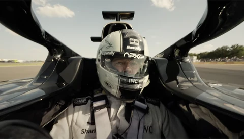 Driver in racing helmet and suit inside open-cockpit race car, captured from above on runway.