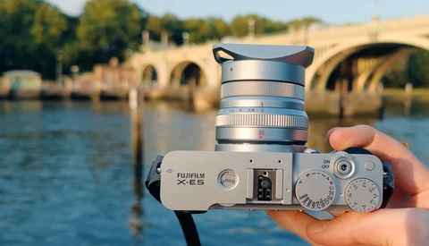 Fujifilm X-E5 camera with silver lens held in hand over a river and stone bridge.