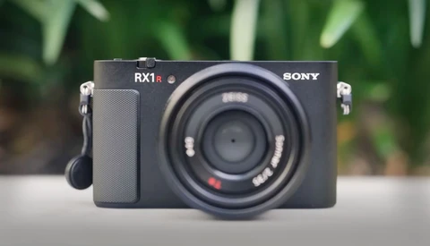 Sony RX1R compact camera with fixed lens, displayed front-facing against blurred green foliage background.