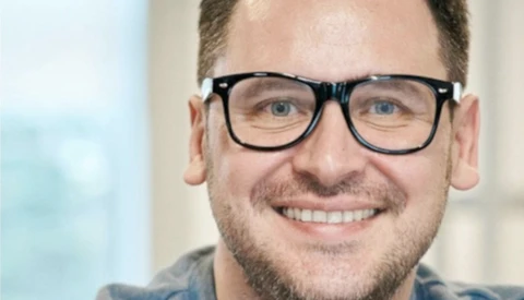 Man with glasses smiling at camera in bright indoor setting.