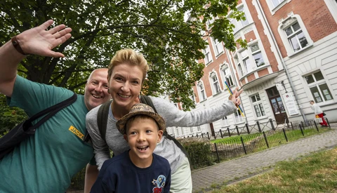 Three people posing together outdoors with arms raised, smiling at the camera in front of a brick building.