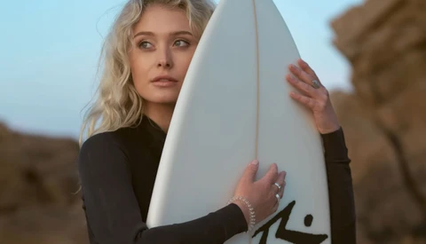 Blonde woman in black wetsuit holding white surfboard at rocky coastline.