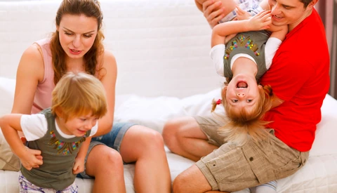 Family of four playing together on a white bed, with parents and children laughing and playfully interacting.