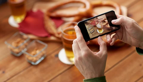 Person holding a smartphone capturing a flat lay composition of appetizers and drinks on a wooden table.