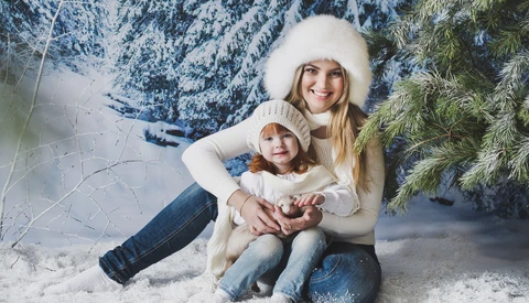 Woman and child sitting together in snow wearing matching cream-colored winter hats and sweaters.