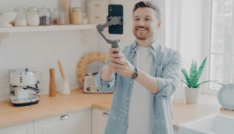 Man holding smartphone on gimbal stabilizer in bright kitchen with white cabinetry.