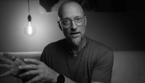Man with glasses wearing a dark t-shirt gestures while speaking, lit by a hanging incandescent bulb against a dark background.