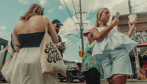 Woman holding a canvas tote bag with script lettering walks down a sunny street with other pedestrians.