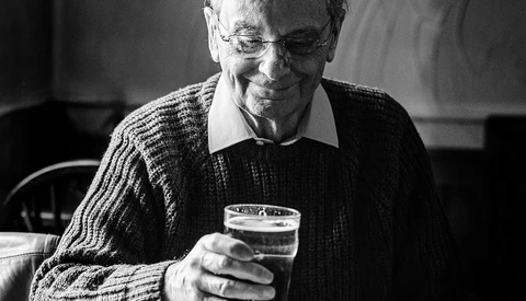 Older man wearing glasses and a cardigan holding a glass of water, looking down with a gentle expression.