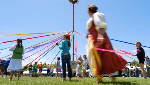 Children and adults holding colorful ribbons attached to a tall maypole during an outdoor spring celebration on a sunny day.