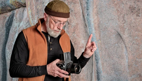 Older man with beard and glasses holding a vintage film camera against a textured stone wall.
