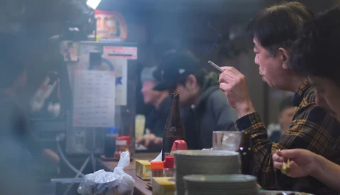 Woman holding chopsticks over a bowl in a smoky kitchen environment with other diners visible in background.