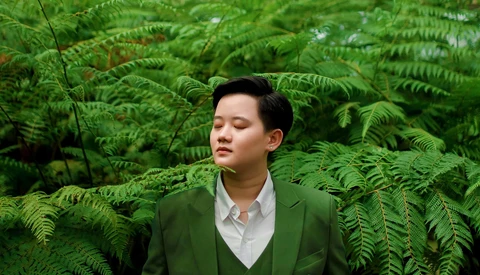 Young man in green blazer surrounded by dense fern foliage, eyes closed in contemplative pose.