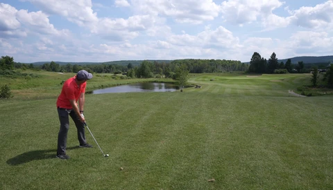 Golfer in red shirt taking a shot on a wide-open fairway with a pond and tree line visible in the distance.