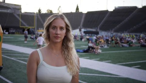 Portrait of a woman with blonde wavy hair on a college football field during an event with crowds in the stadium background.
