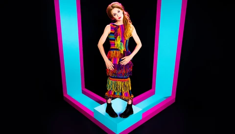 Woman in colorful fringed outfit posed against bright cyan and magenta geometric neon lines.