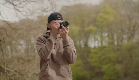 Photographer wearing a cap and tan jacket holding binoculars to their eyes in a wooded outdoor setting.