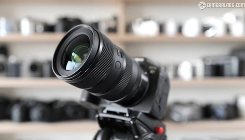 Black zoom lens mounted on camera body positioned on tripod against blurred equipment shelves.