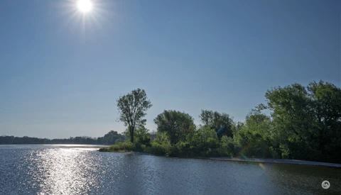 Bright sunlit river landscape with trees lining the far bank under clear blue sky.