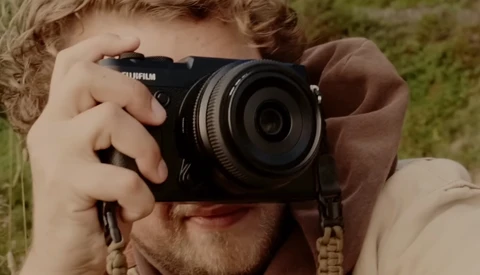 Photographer holding a Fujifilm mirrorless camera up to his face to compose a shot outdoors.