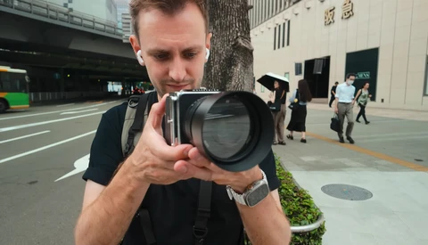 Photographer holding a large telephoto lens while looking through the viewfinder on an urban street.