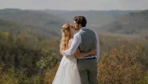 Couple in wedding attire kissing on a hillside overlooking forested valley.