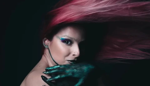 Woman with burgundy hair and teal eyeshadow looks over her shoulder with teal-painted hands near her face against dark background.