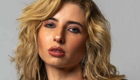 Portrait of a woman with shoulder-length wavy blonde hair against a neutral gray background.