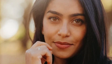 Woman with long dark hair resting her chin on her hand, gazing directly at camera with warm expression.