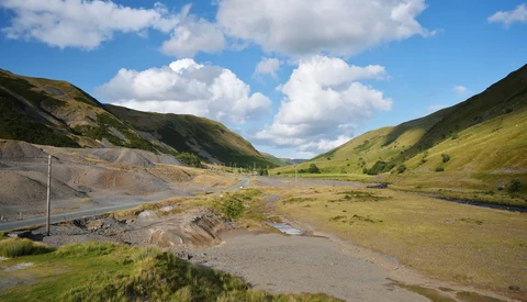 Mountain valley with grassy slopes, exposed earth, and a small stream under blue sky with white clouds.