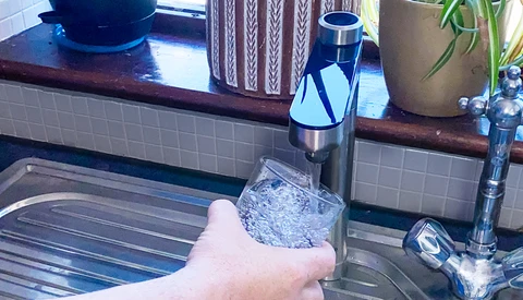 Hand holding a glass of water from a kitchen faucet with aerator visible.