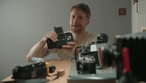 Photographer examining a vintage film camera while surrounded by other classic cameras on a wooden desk.