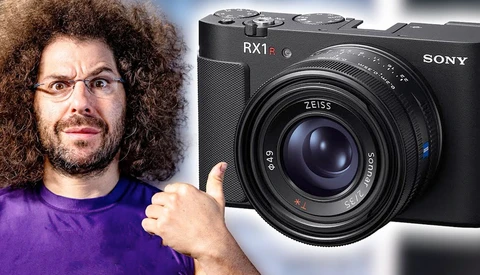 Man in purple shirt with curly hair and glasses gesturing toward a Sony RX1 II compact camera.