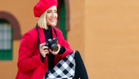 Woman in red blazer and beret holding a mirrorless camera against a warm tan wall.