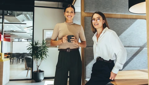Two women smiling in a modern office space, one holding a phone.