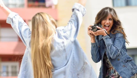 Woman with camera photographing friend with raised arms outdoors.