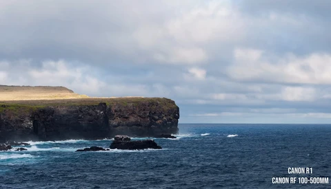 Dramatic coastal cliffs with turquoise waves crashing against dark basalt rock formations under a partly cloudy sky.