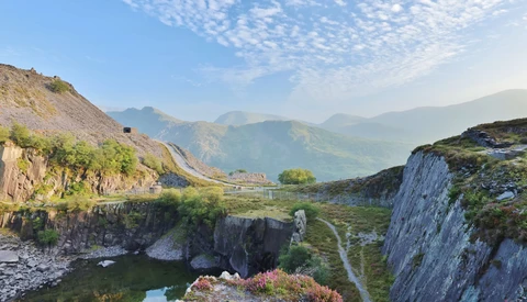Alpine valley landscape with layered mountains, rocky cliffs, and winding river below clear sky.