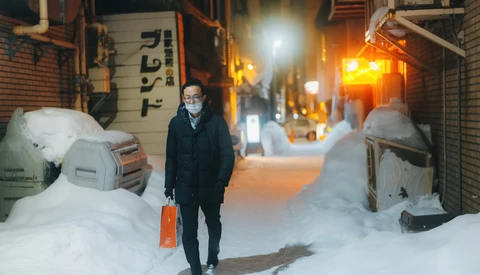 Person standing in snowy alley at night holding red shopping bag, wearing mask and dark coat.