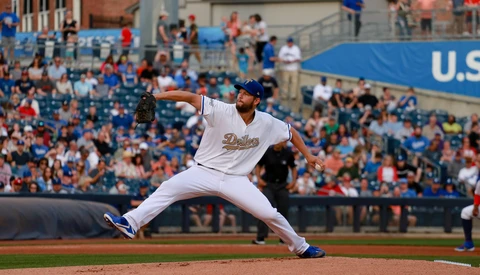 Baseball pitcher in mid-delivery, stretching across the mound in white uniform during a daytime game.