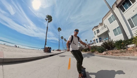 Person riding an electric skateboard on a coastal path with palm trees and beach buildings visible.