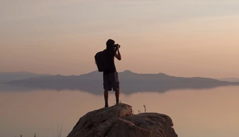 Silhouetted photographer standing on a rock, holding camera to face, overlooking a misty lake and distant mountains at dawn.