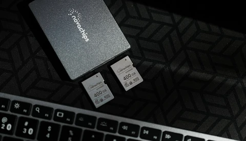 Two SD memory cards next to a dark gray storage case on a patterned surface beside a keyboard.